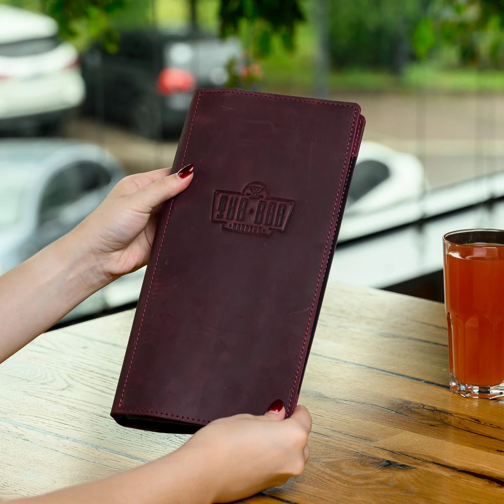 Person holding a maroon leather wallet with a logo, next to a glass of juice on a wooden table.