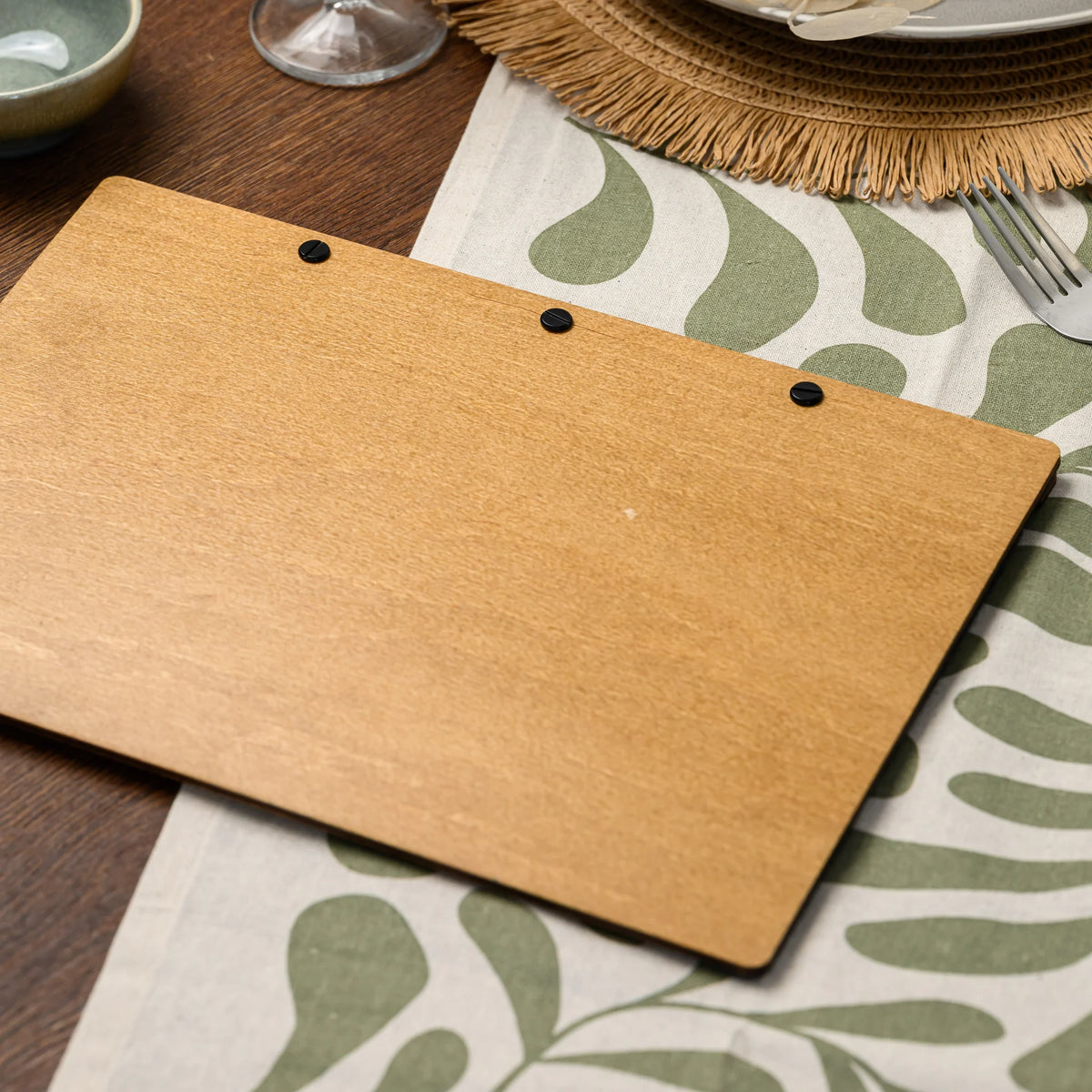 The back panel of a wood-backed felt menu folder resting on a table beside a green leaf-patterned napkin 