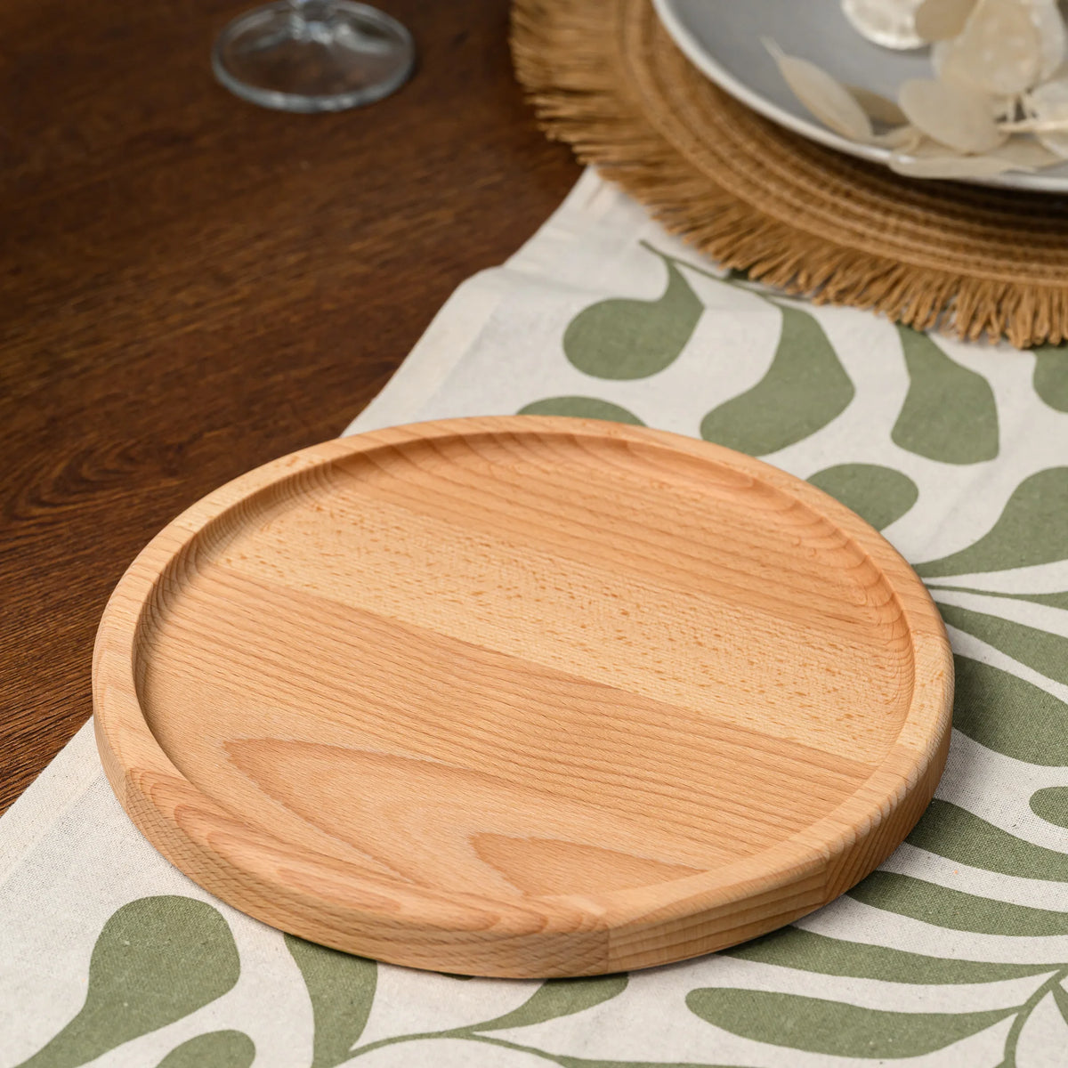 Overhead angled view of an empty round beech wood serving plate resting on a leaf-patterned tablecloth