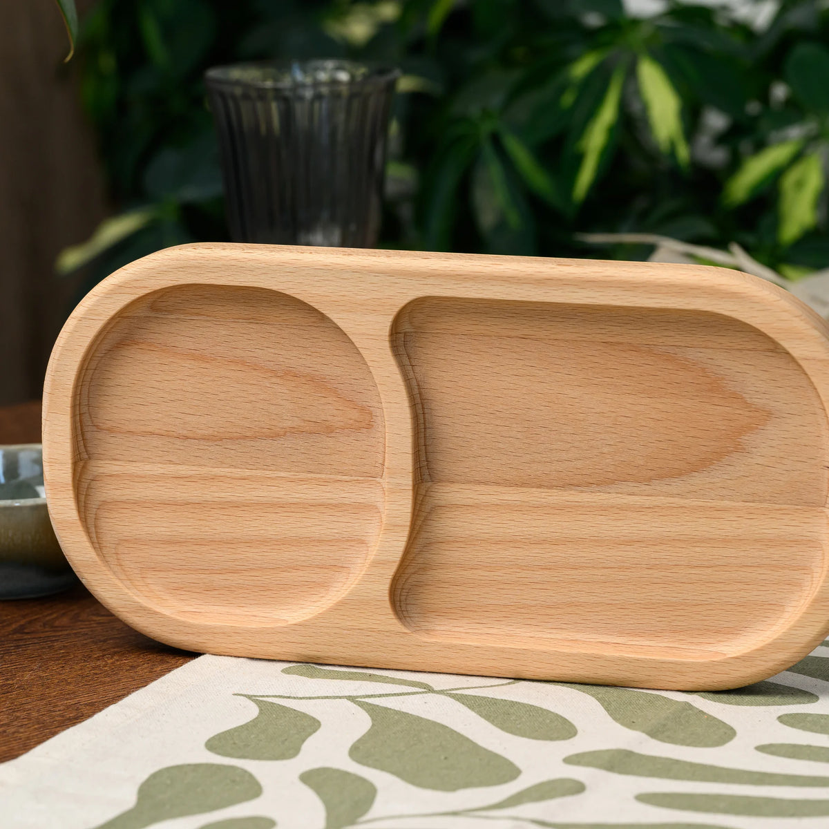 Close view of an empty two-compartment beech wood serving board resting among green foliage in the background