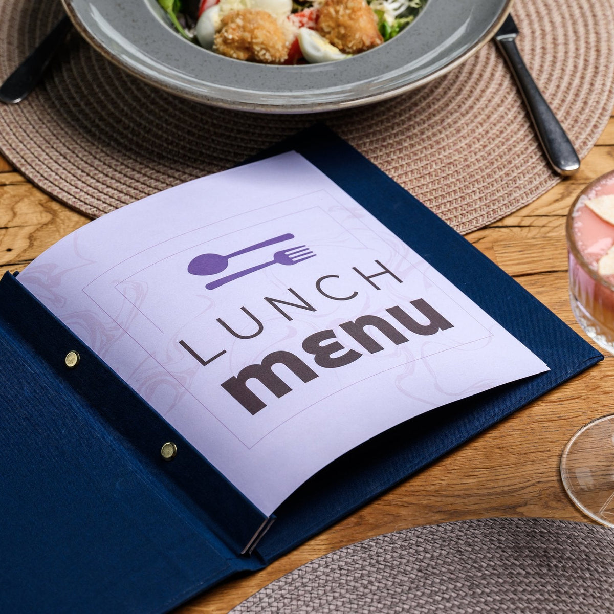 Wide open navy textile menu folder with a white lunch menu page beside a plate of food and a pen on placemat