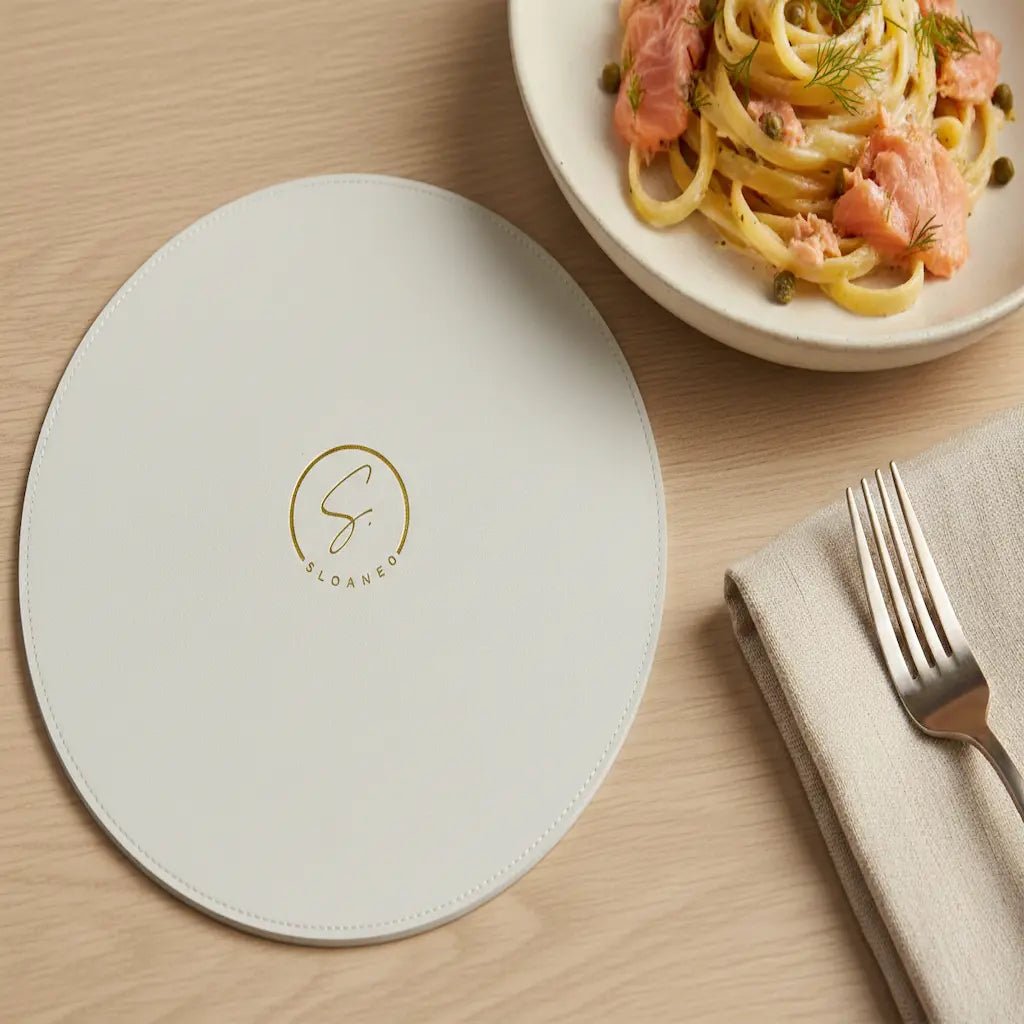 Round white placemat with a logo on a wooden table with pasta dish and cutlery.