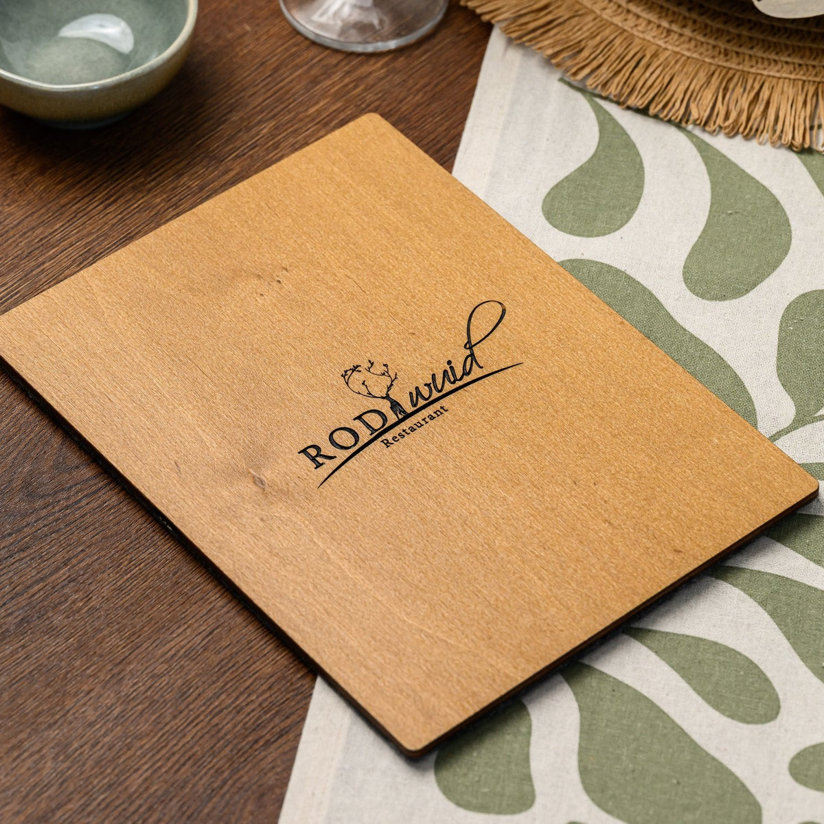 The plywood back of a felt-front menu clipboard, resting flat on a table with its engraved logo visible 