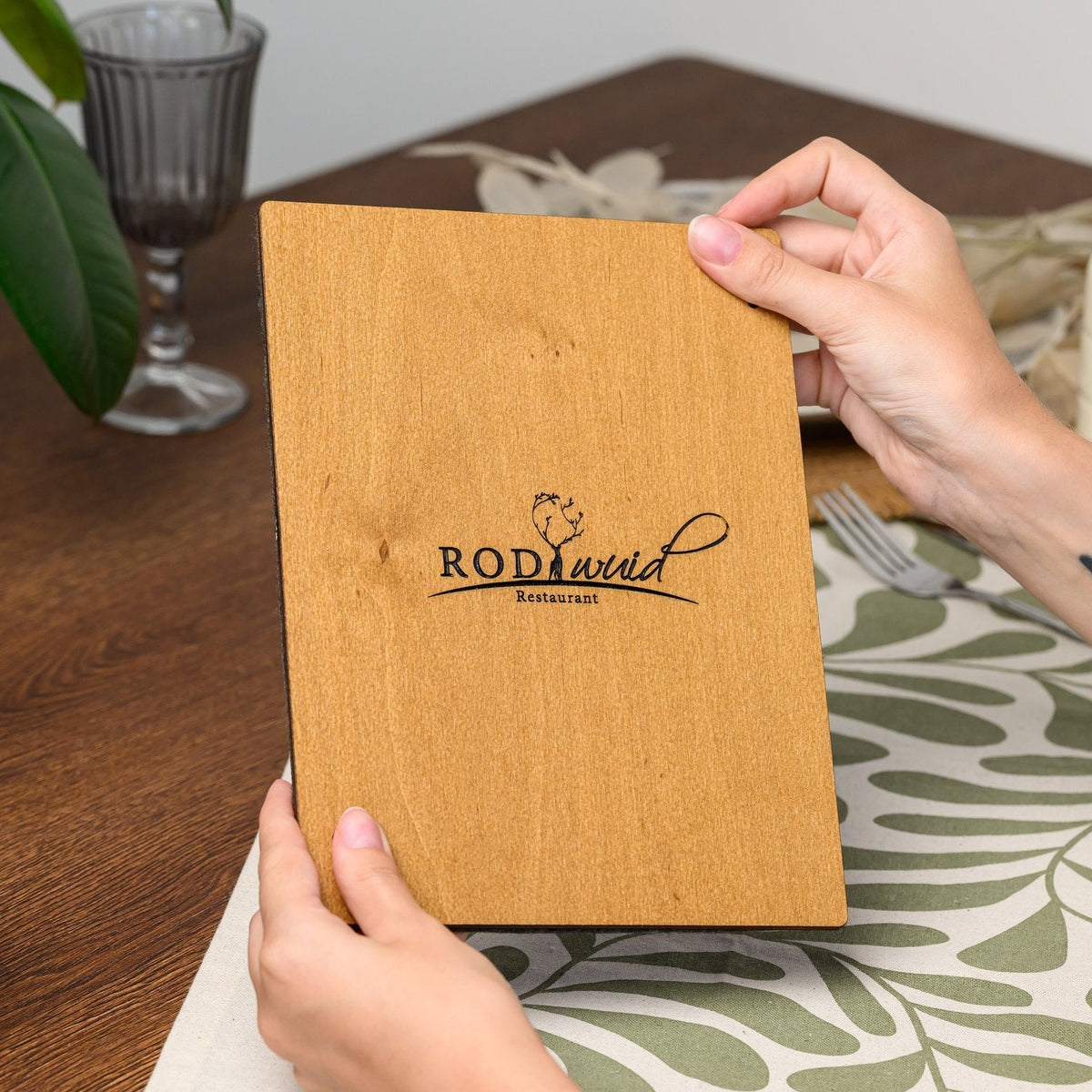 A hand holding the reverse side of a menu clipboard, showing an engraved logo on the plywood back panel 