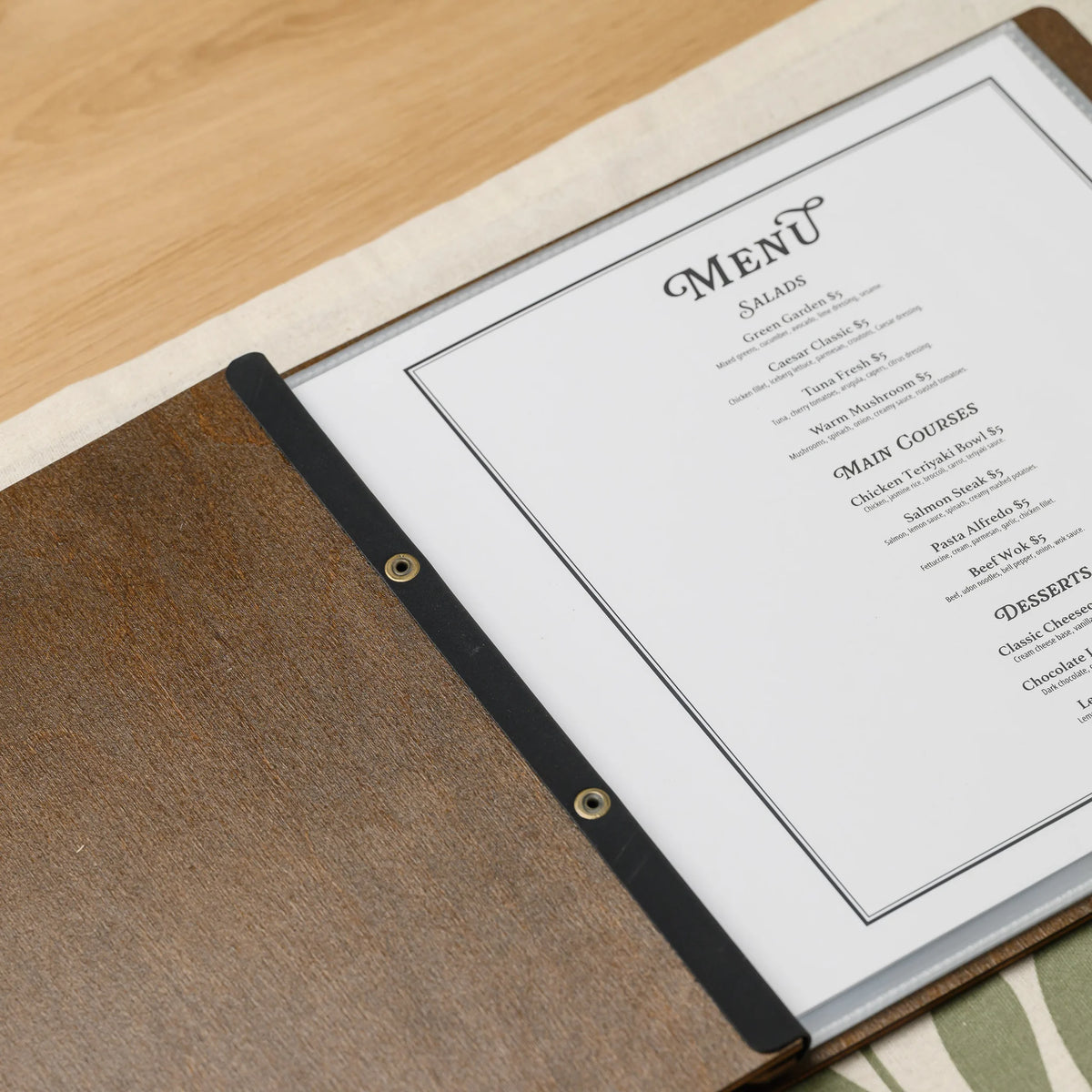 Open walnut plywood menu cover with a printed menu in a clear sleeve held by a black leather strap with eyelets