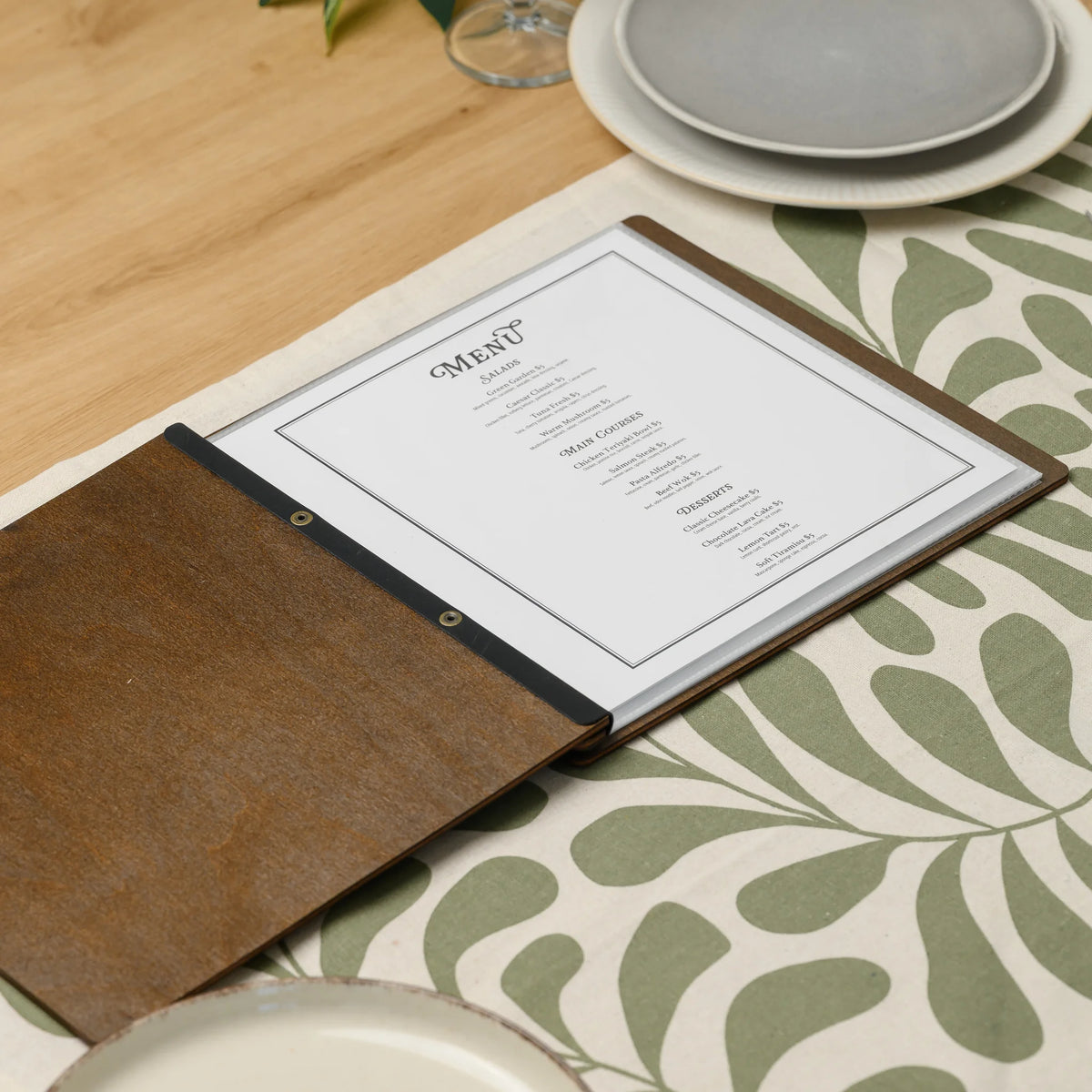Open walnut plywood menu cover on a set table with a printed menu in a clear sleeve and a black leather strap