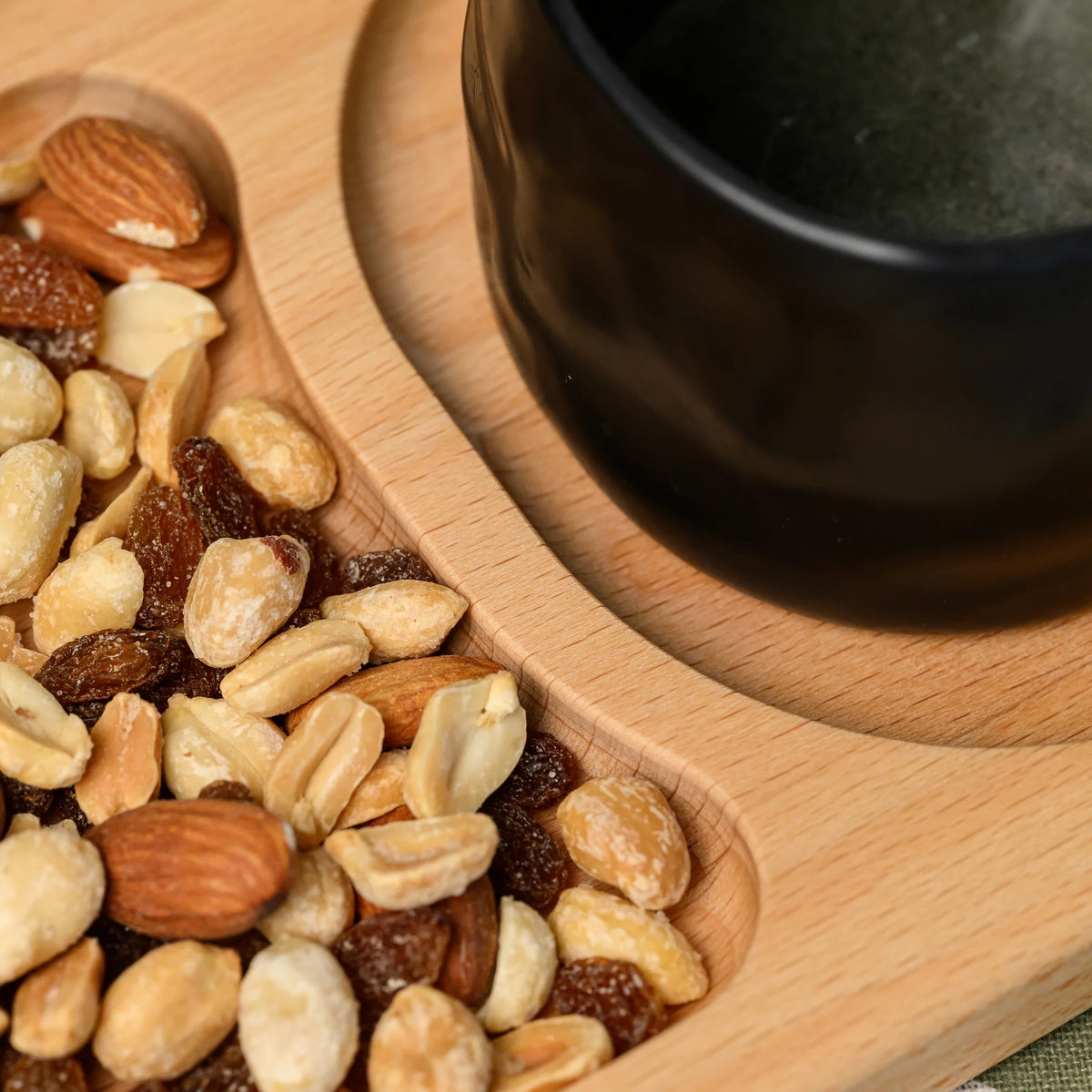 Close-up of mixed nuts and raisins beside a black ceramic mug in the beech wood board's oval compartment