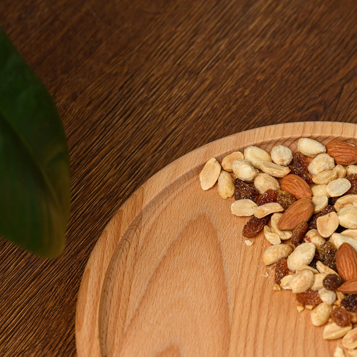 Top-down close-up of the beech wood plate's corner with mixed nuts and raisins, green leaf overhead