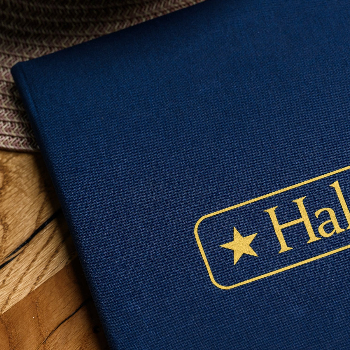Closed navy textile menu folder standing upright with a gold star-badge logo partly visible on a placemat
