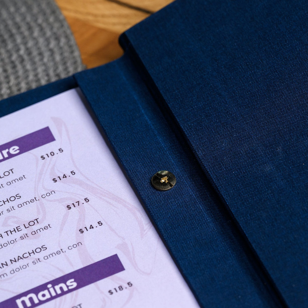 Open navy textile menu folder shown from the side with a purple menu page and a gold snap fastener on wood