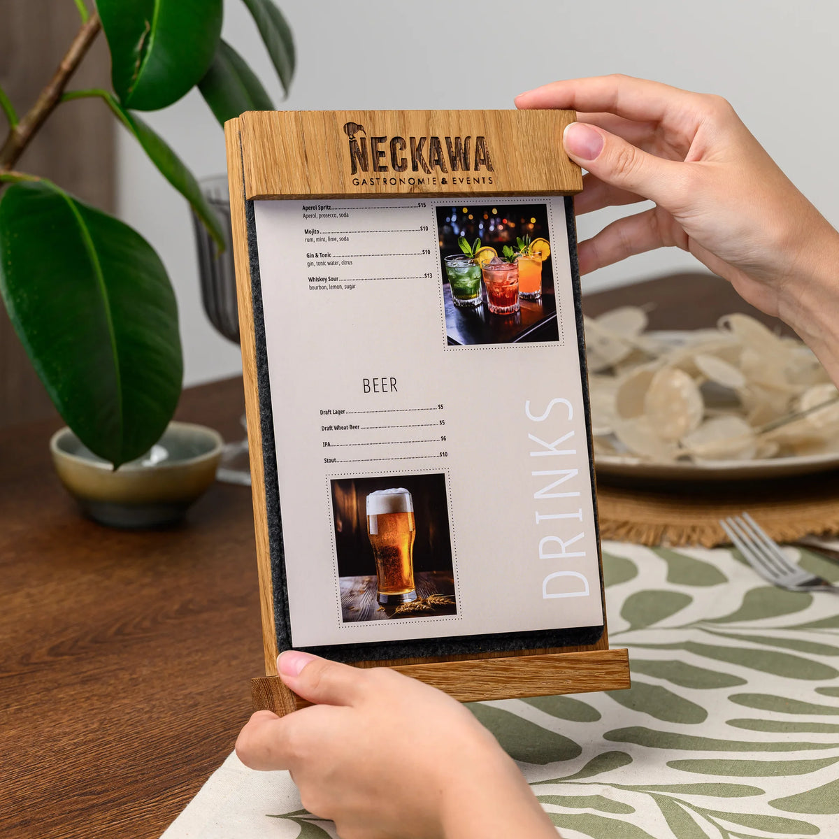 Hands holding open a table tent, showing the engraved oak bar and a printed drinks menu page held inside 