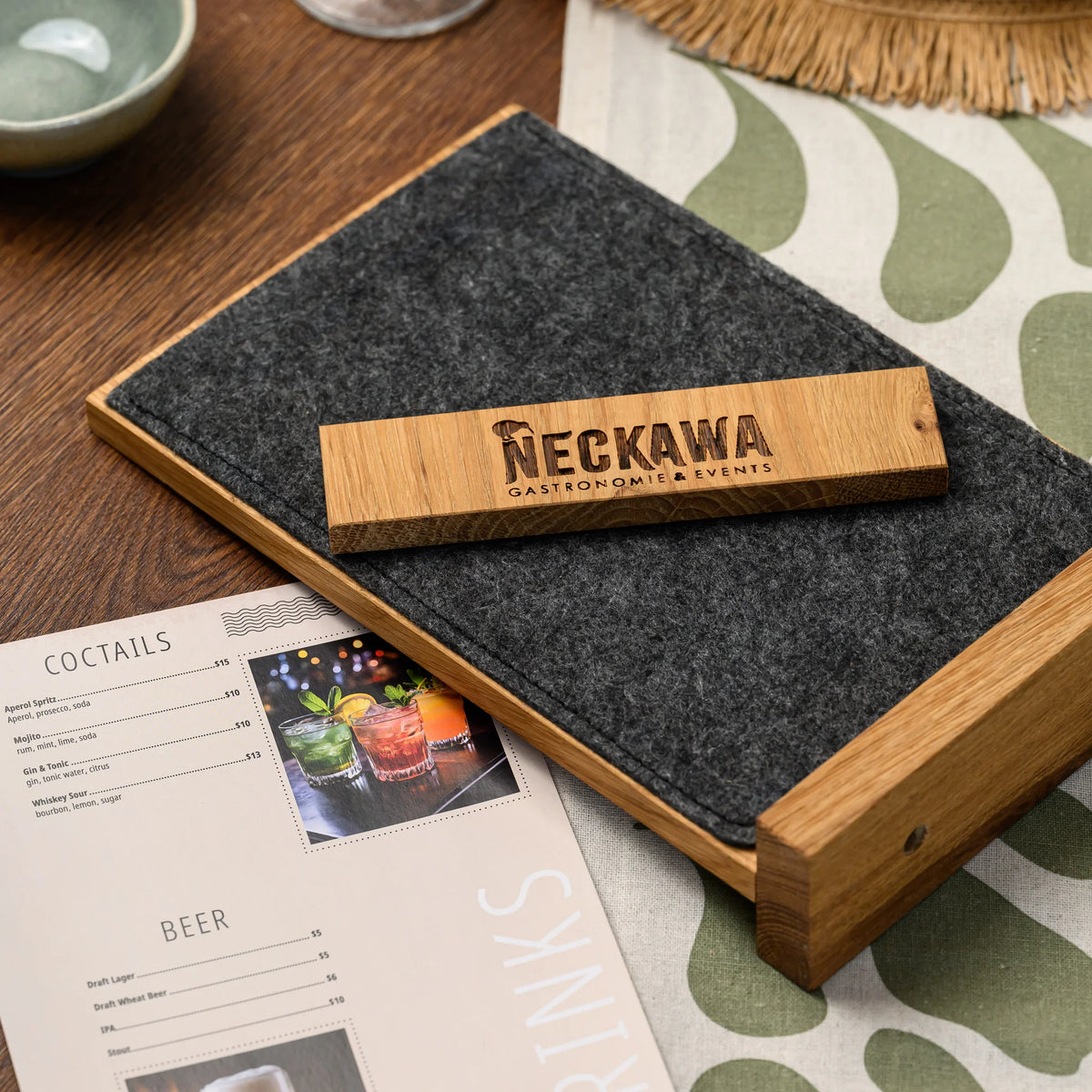 A close-up of an engraved oak bar lying flat on a felt panel beside a printed cocktail menu photograph 