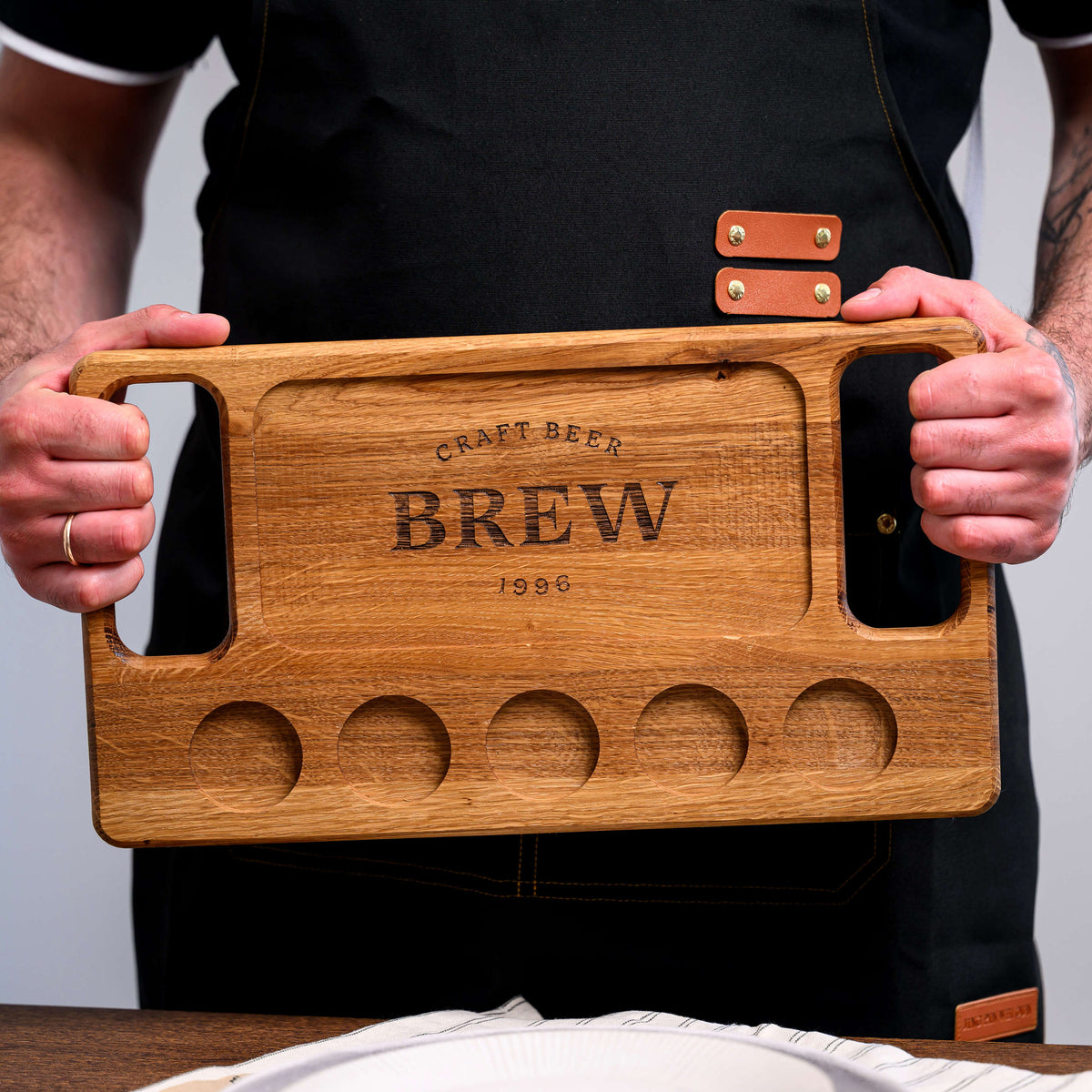 Rectangular oak beer flight board held in black apron with both handles, showing Craft Beer Brew 1996 engraving