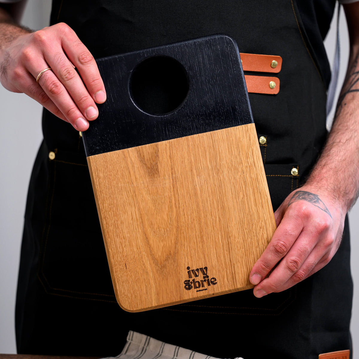 Two-tone oak snack board with black painted handle held in dark apron, showing hanging hole and Ivy and Brie mark