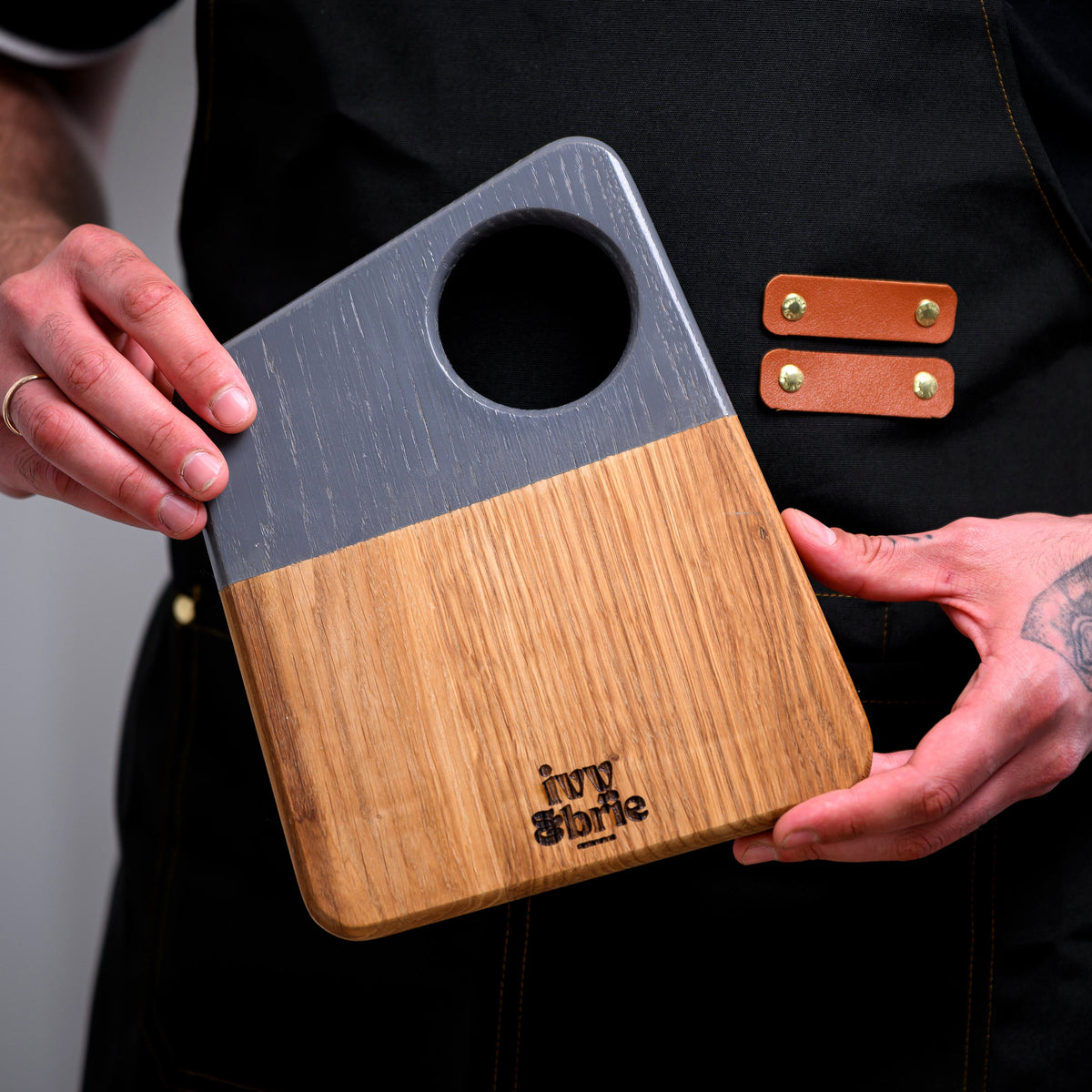 Two-tone oak snack board with grey-dipped painted handle held in dark apron, showing hanging hole and Ivy and Brie mark