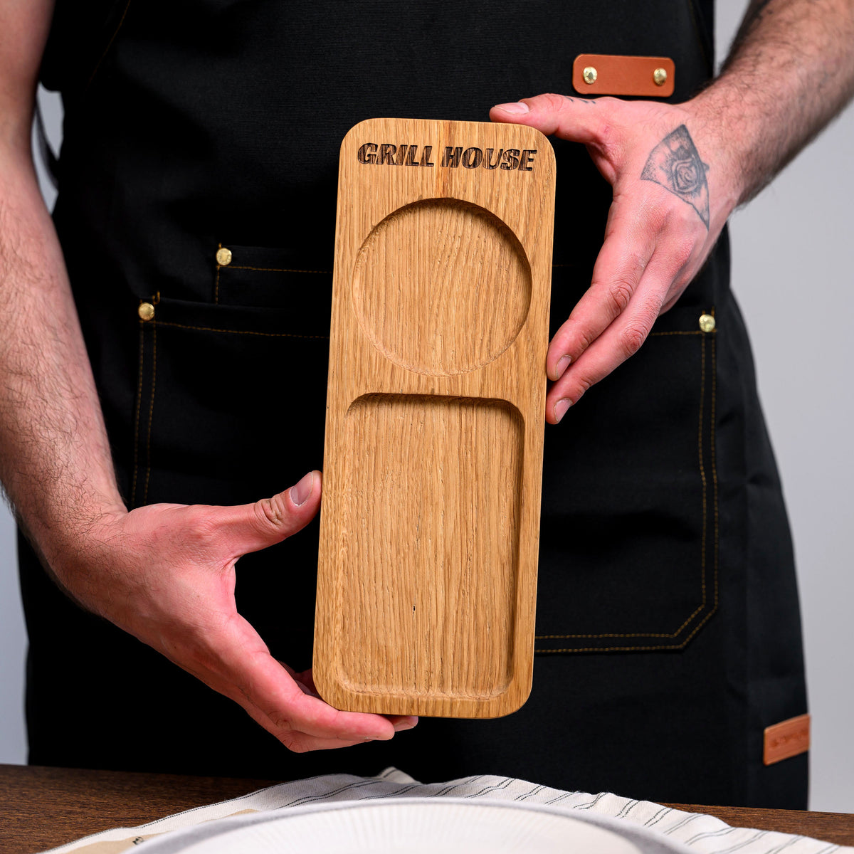 Oak two-section serving board with GRILL HOUSE engraving held in dark apron, showing goblet recess and snack tray