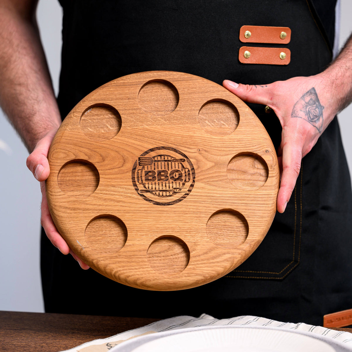 Round oak BBQ serving board held in front of a black apron, showing scale, full form and central engraved grill emblem