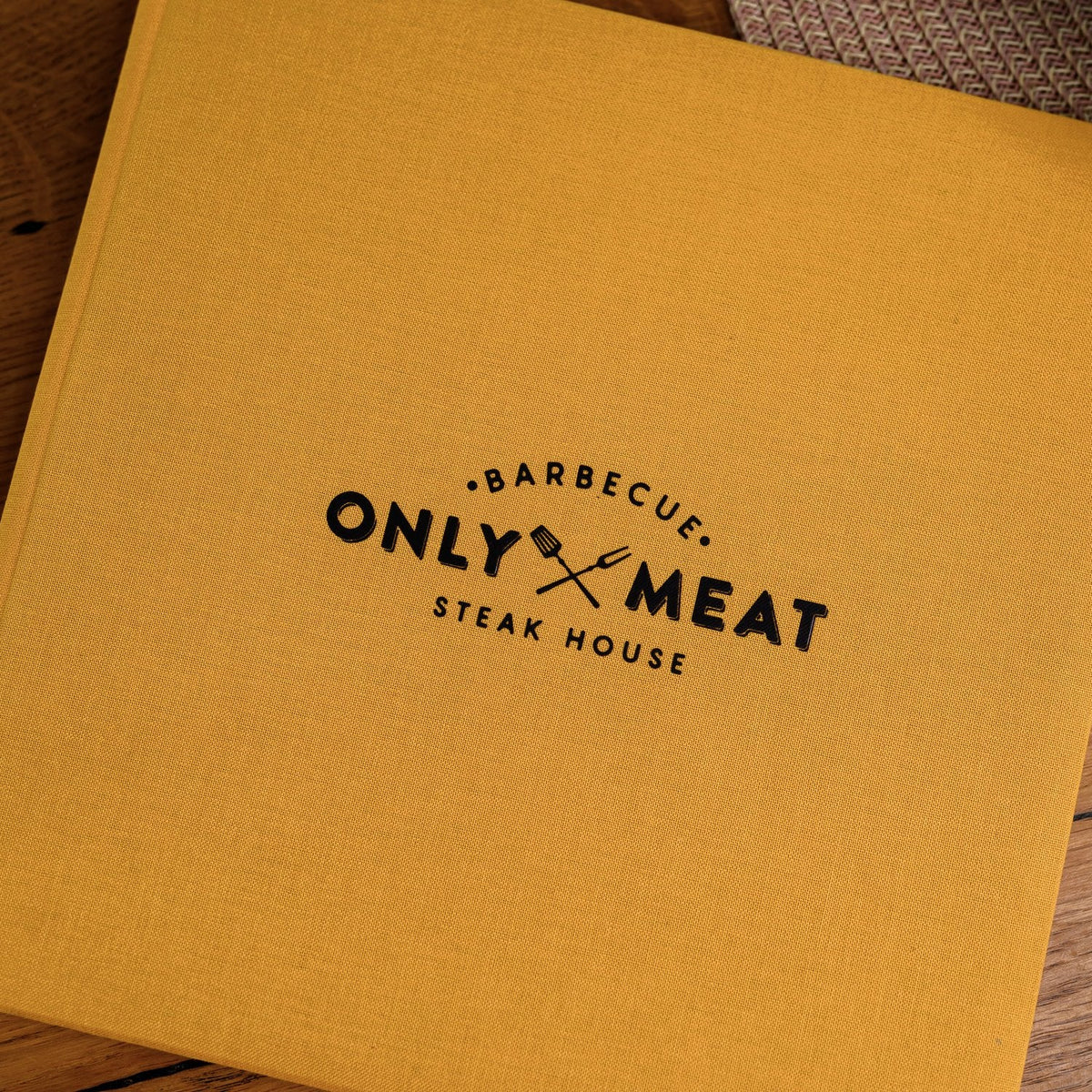 Closed mustard-yellow textile menu folder standing upright with a centered black logo on a wooden table