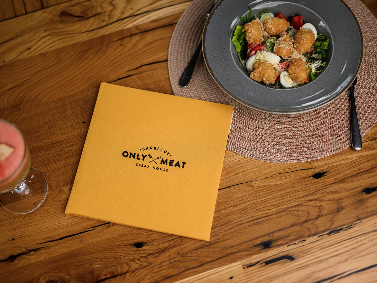 Small closed mustard-yellow textile menu folder with a black logo on a wooden table, blurred plate nearby