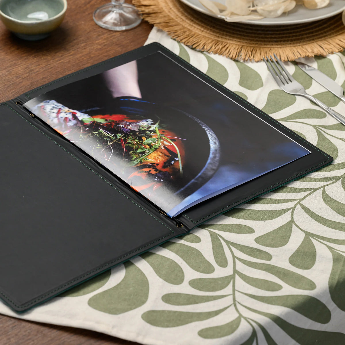 Open green leather menu folder lying flat on a set table with a food photo menu page and black inner lining