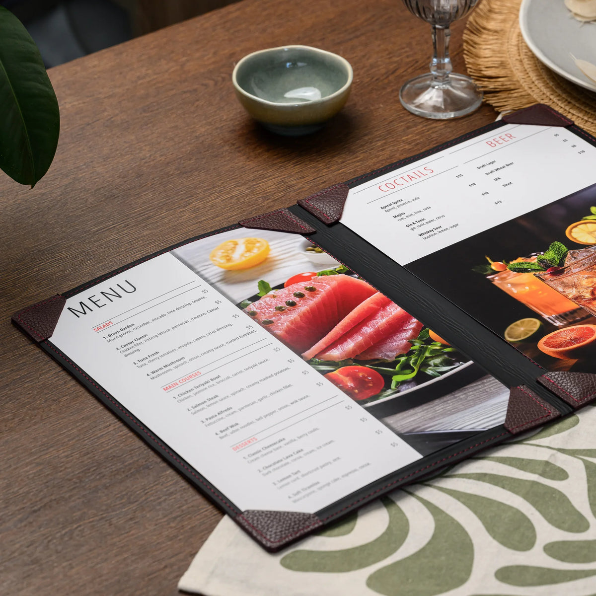 Open bordeaux leather menu cover on a set table with two printed sheets held flat by stitched leather corners