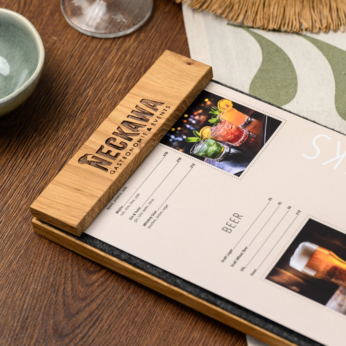 The engraved oak bar of a table tent resting on a charcoal felt panel next to a printed drinks menu page 