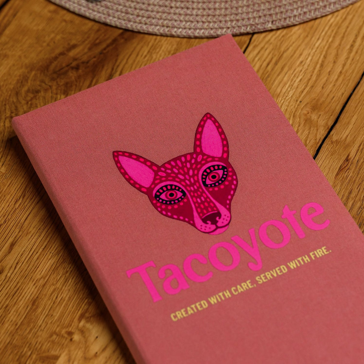 Closed coral-pink textile menu folder standing upright with a fox graphic on a wooden restaurant table