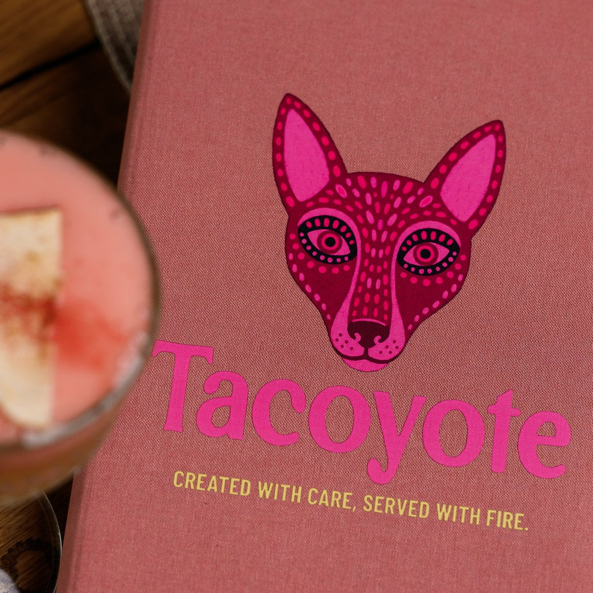 Close-up of a closed coral-pink textile menu folder with a fox graphic beside blurred citrus and melon slices
