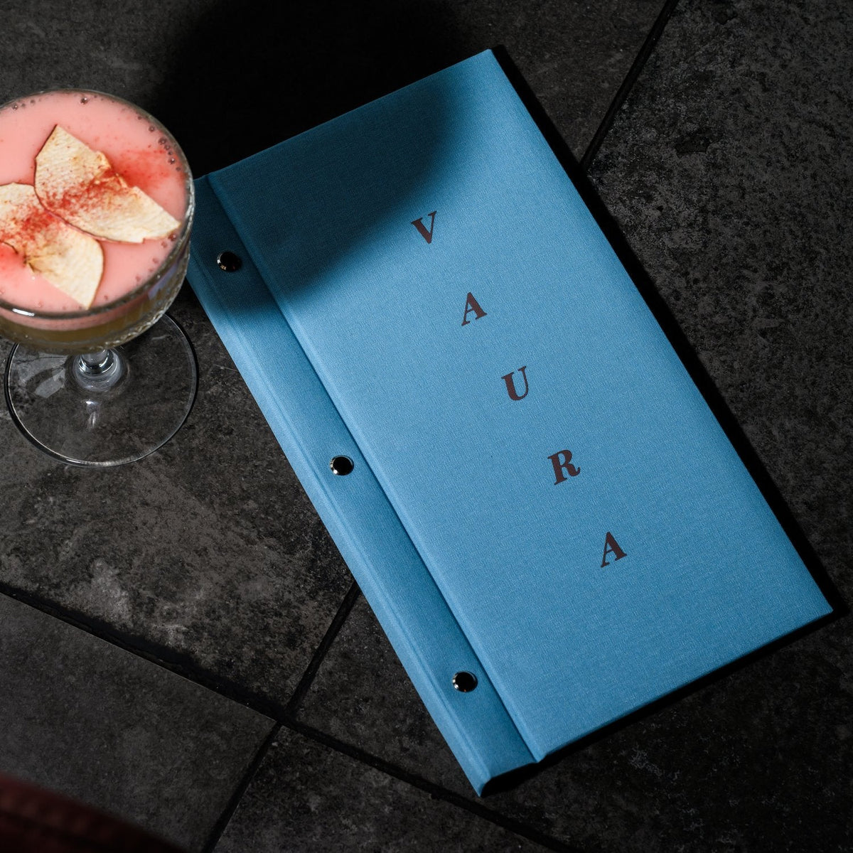 Closed light-blue woven-textile menu folder with vertical spaced lettering lying on dark tile beside a cocktail