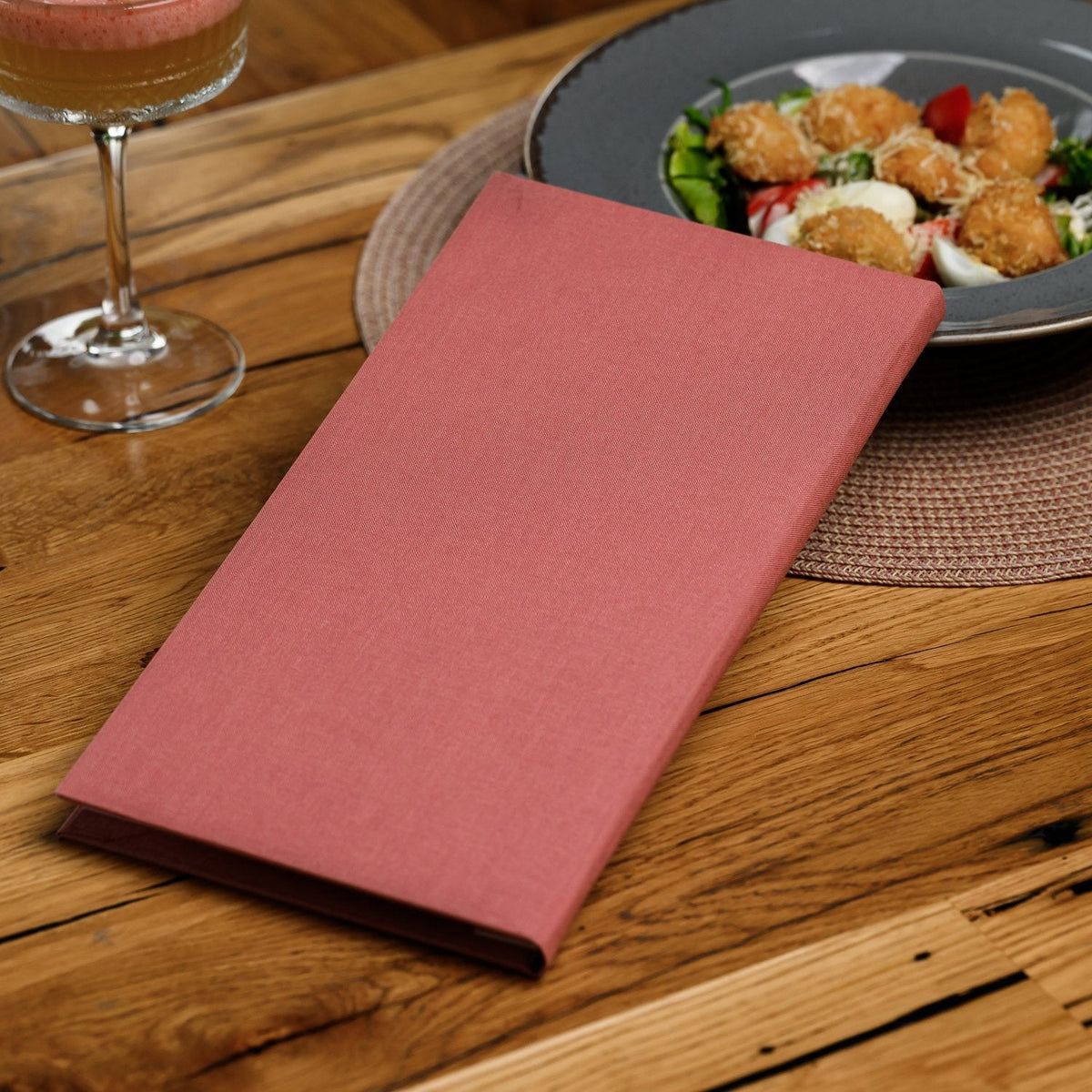 Closed coral-pink textile menu folder lying diagonally beside a wine glass and salad plate on wood table