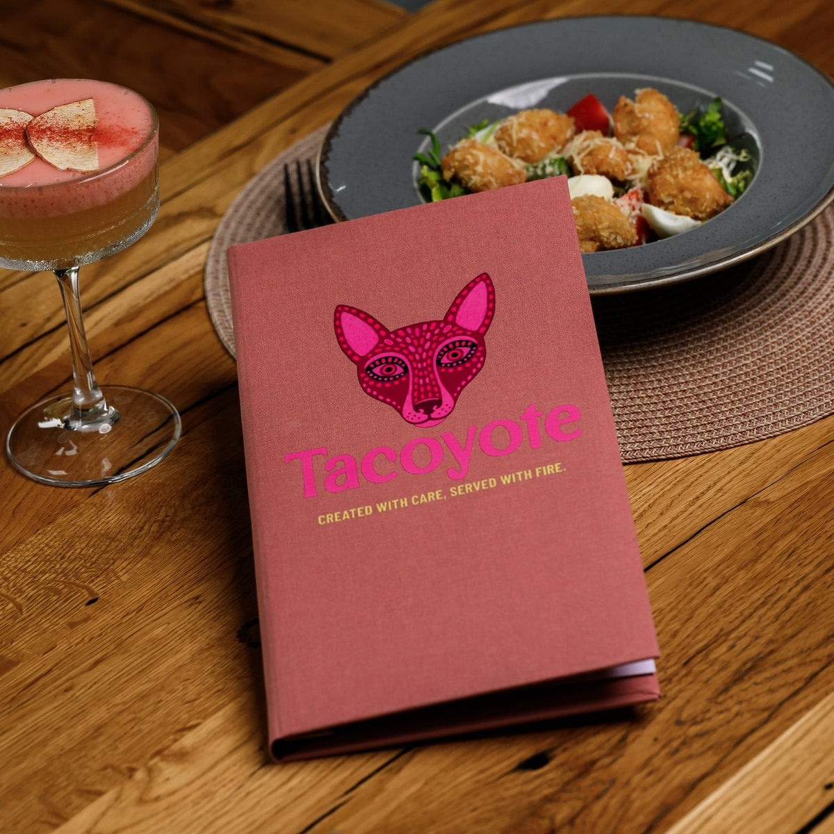 Small closed coral-pink textile menu folder standing beside a cocktail and salad plate on a woven placemat