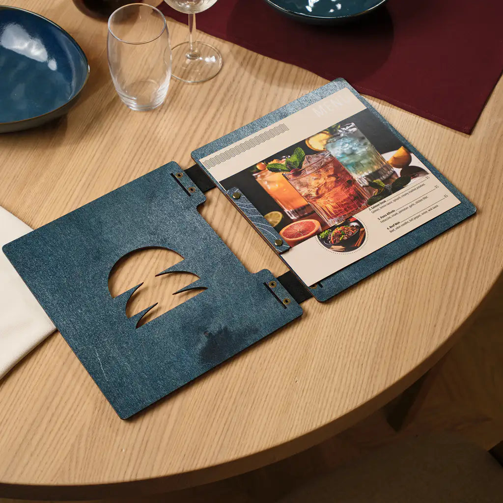 Denim blue plywood square menu folder open on a restaurant table, cocktail menu visible through round cutout