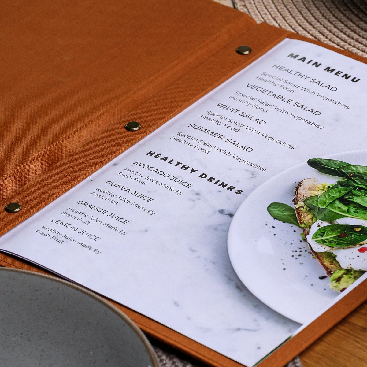 Open rust-orange textile menu folder with a printed menu list beside a plate of fresh green garden salad
