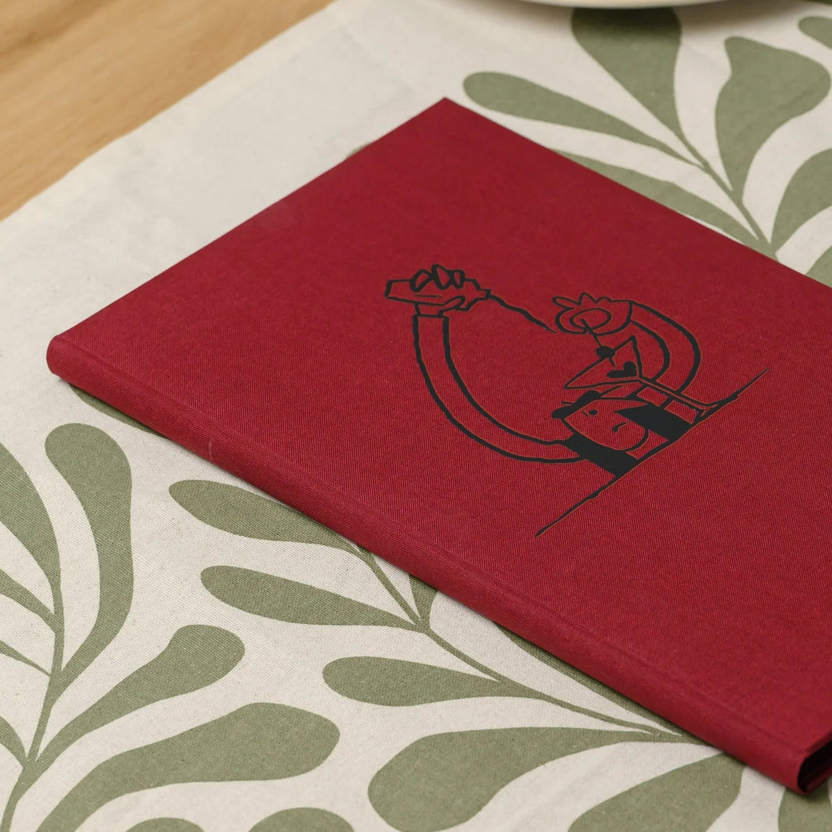 Tall red fabric menu cover with a black printed illustration and a clean front lying on a set restaurant table