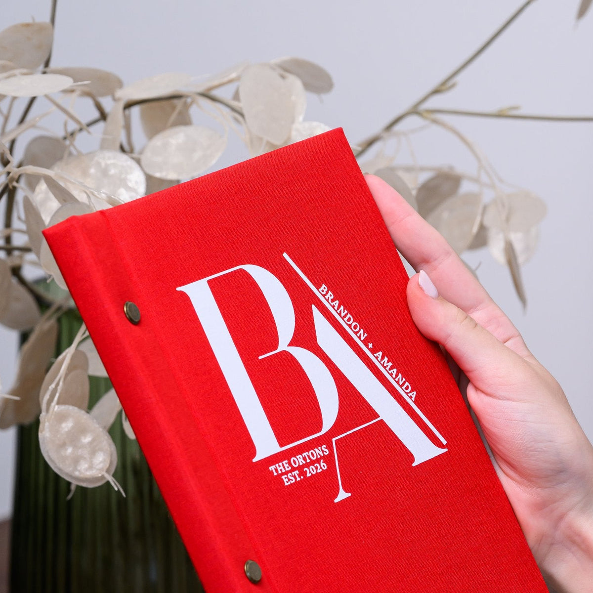 Hand holds a closed red woven-textile menu folder with a bold personalized monogram against dried florals