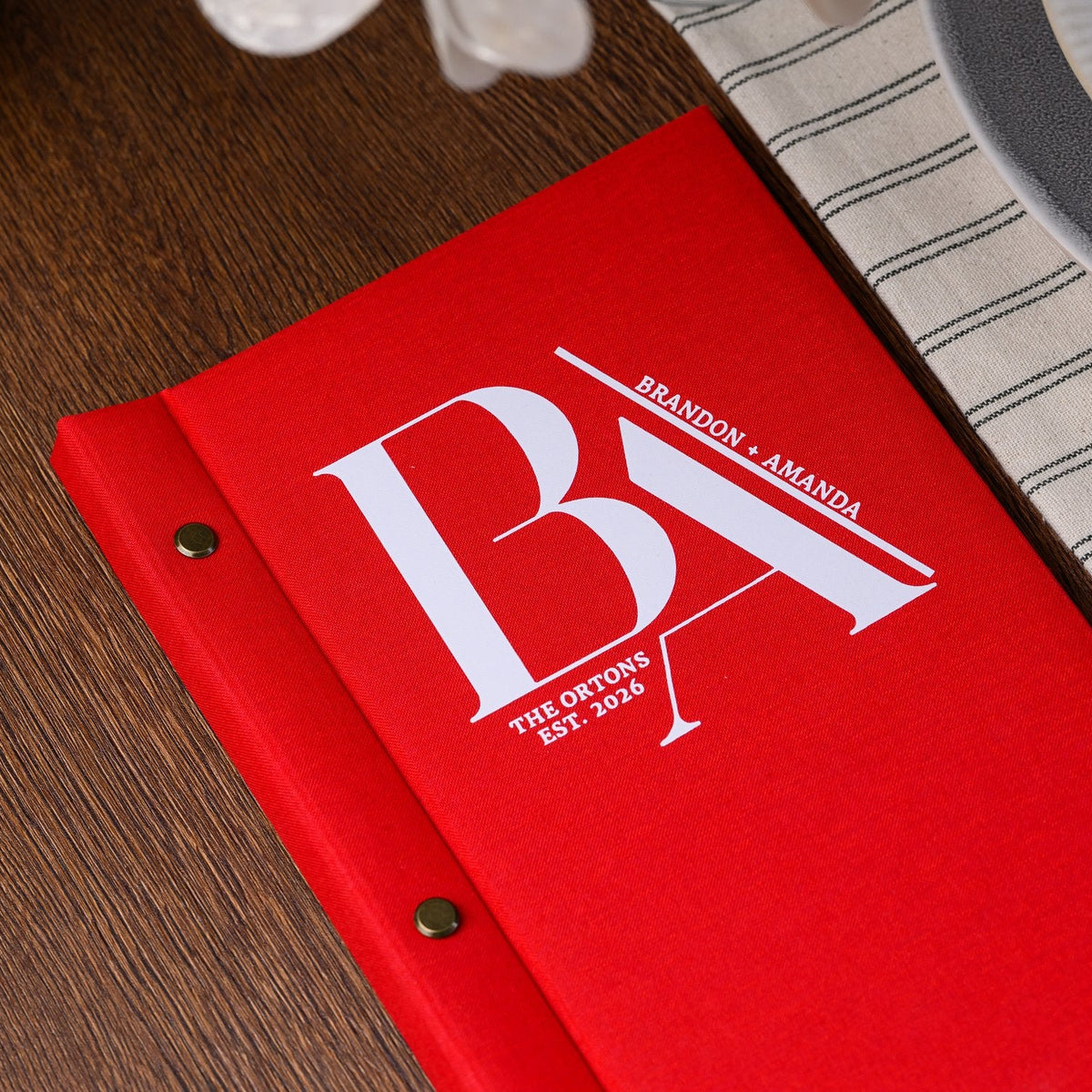 Closed red woven-textile menu folder standing upright with a monogram cover on a striped fabric placemat