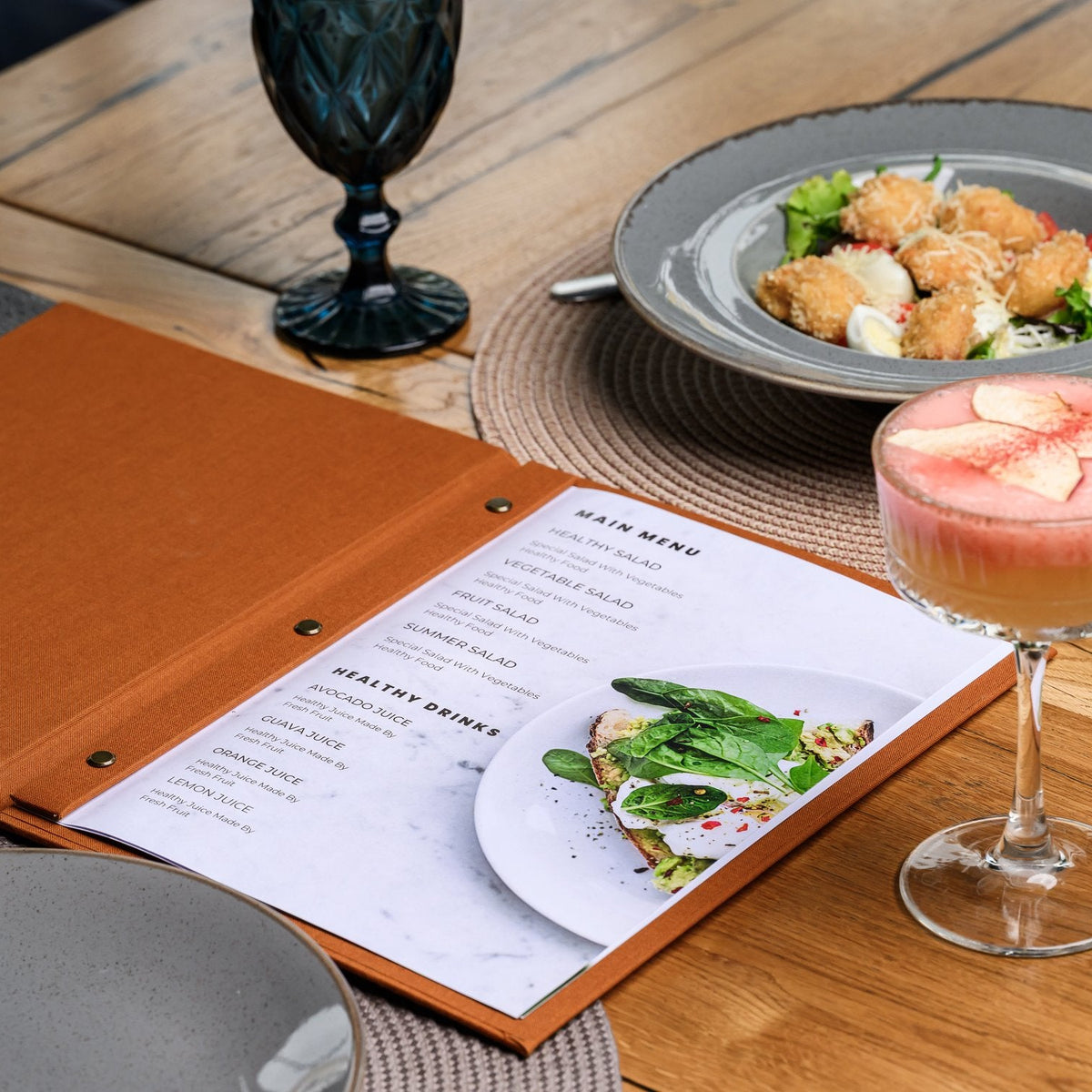 Wide open rust-orange textile menu folder with a printed menu list beside a salad and a garnished cocktail