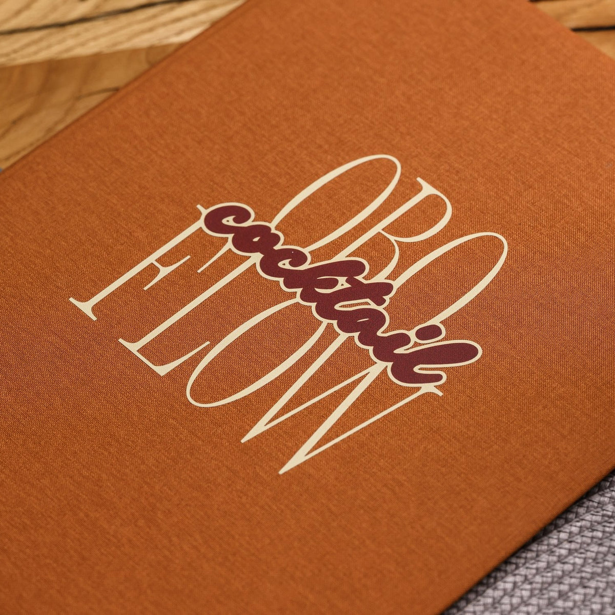 Close-up of a white script logo on the cover of a closed rust-orange textile menu folder on a woven mat