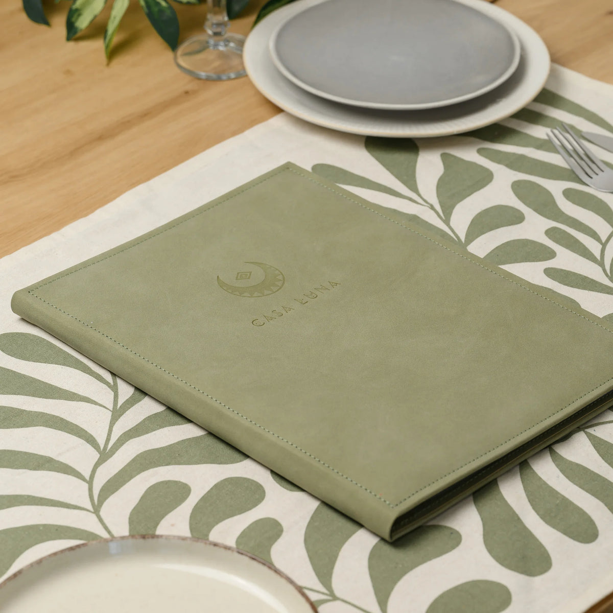 Closed olive Nubuk faux leather menu cover with a blind embossed logo lying on a patterned table runner near plates