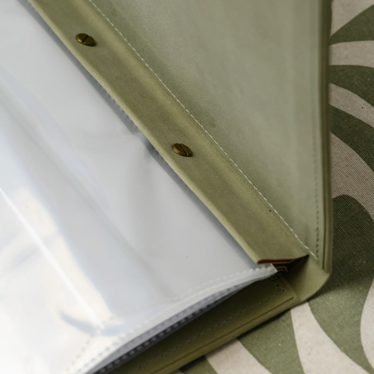 Close-up of slotted brass screws on the plank of an olive Nubuk menu cover with a stack of plastic sleeves beside it