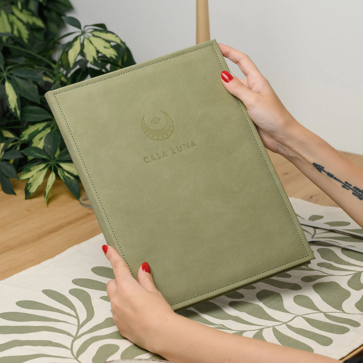 Hands holding a closed olive Nubuk faux leather menu cover with a blind embossed logo above a set restaurant table
