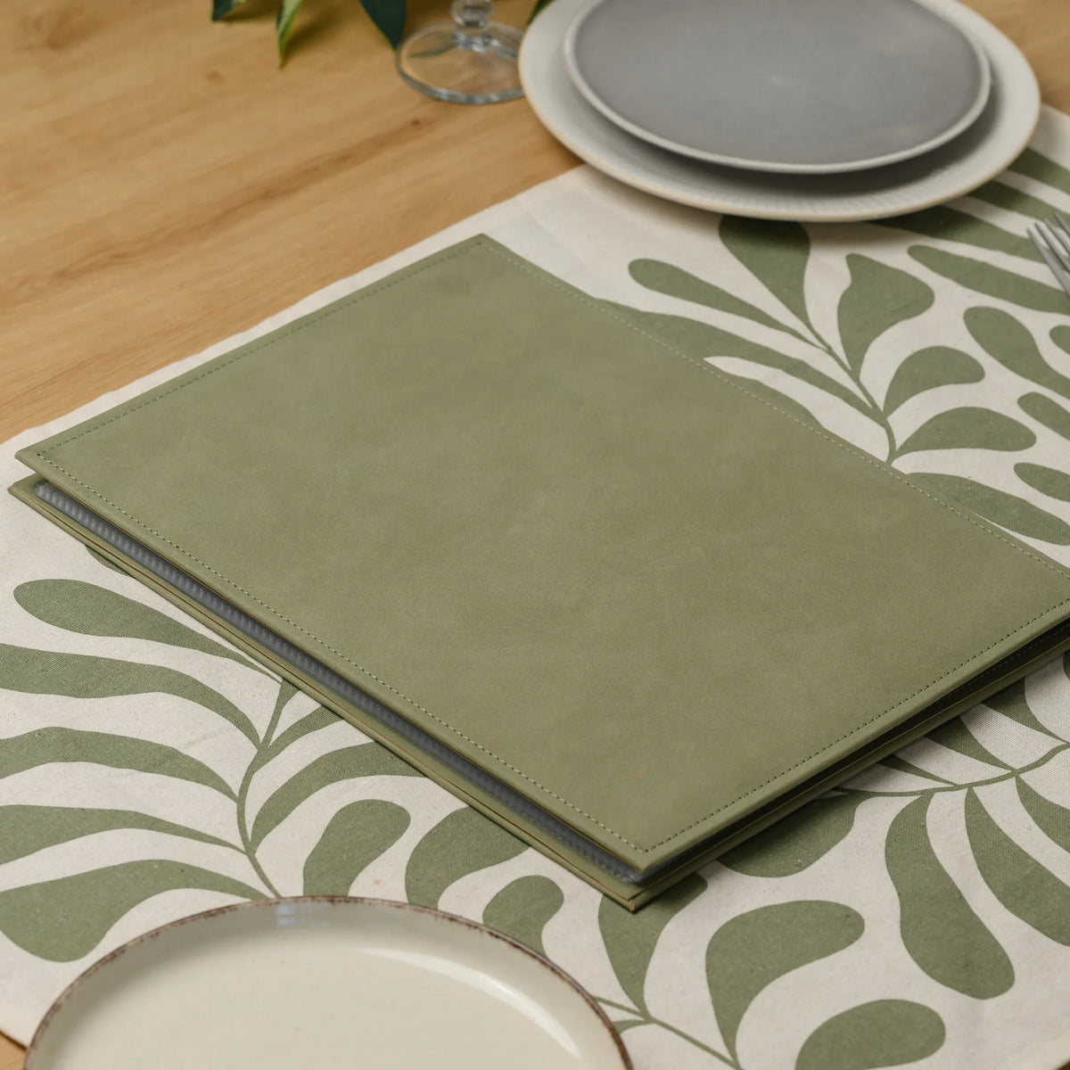Closed olive faux leather menu cover lying on a patterned table runner with the edges of plastic sleeves visible