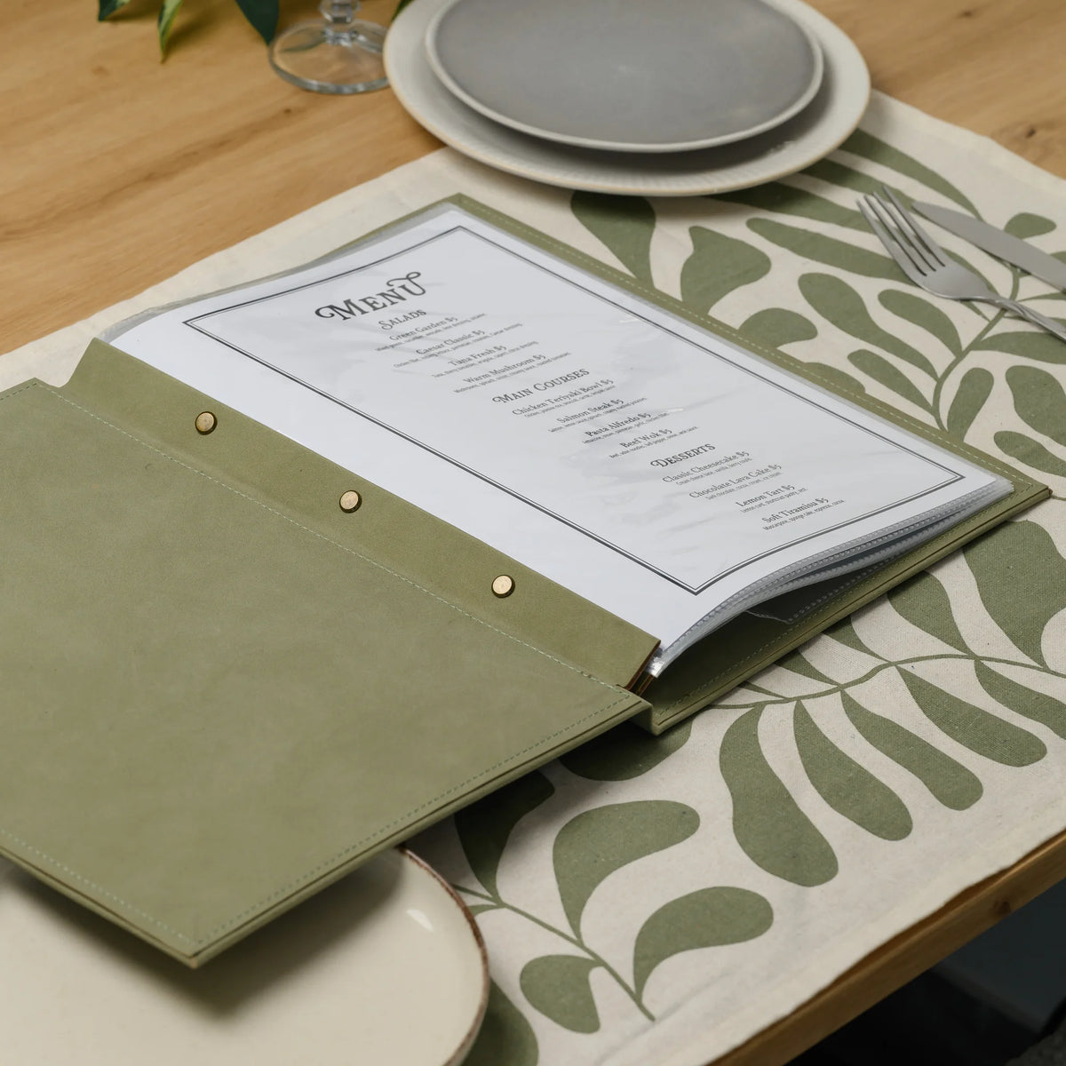Open olive faux leather menu cover on a set table with a menu in a clear sleeve and three brass screws on the plank