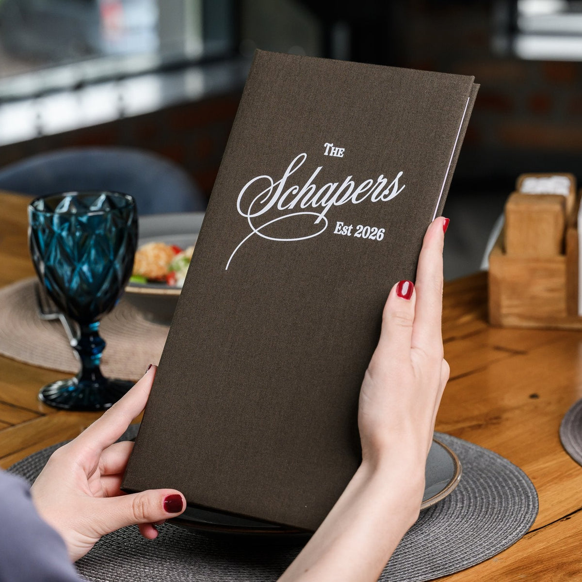 Hand holds a closed espresso-brown textile menu folder standing upright beside a dark blue wine glass