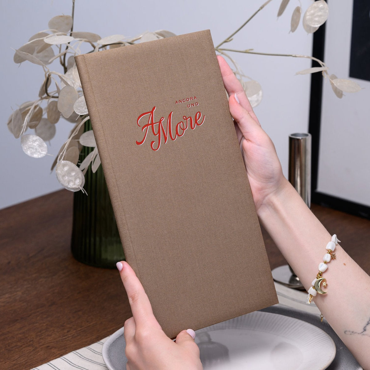 Hand holds an open taupe textile menu folder with a red cursive script logo against blurred dried florals