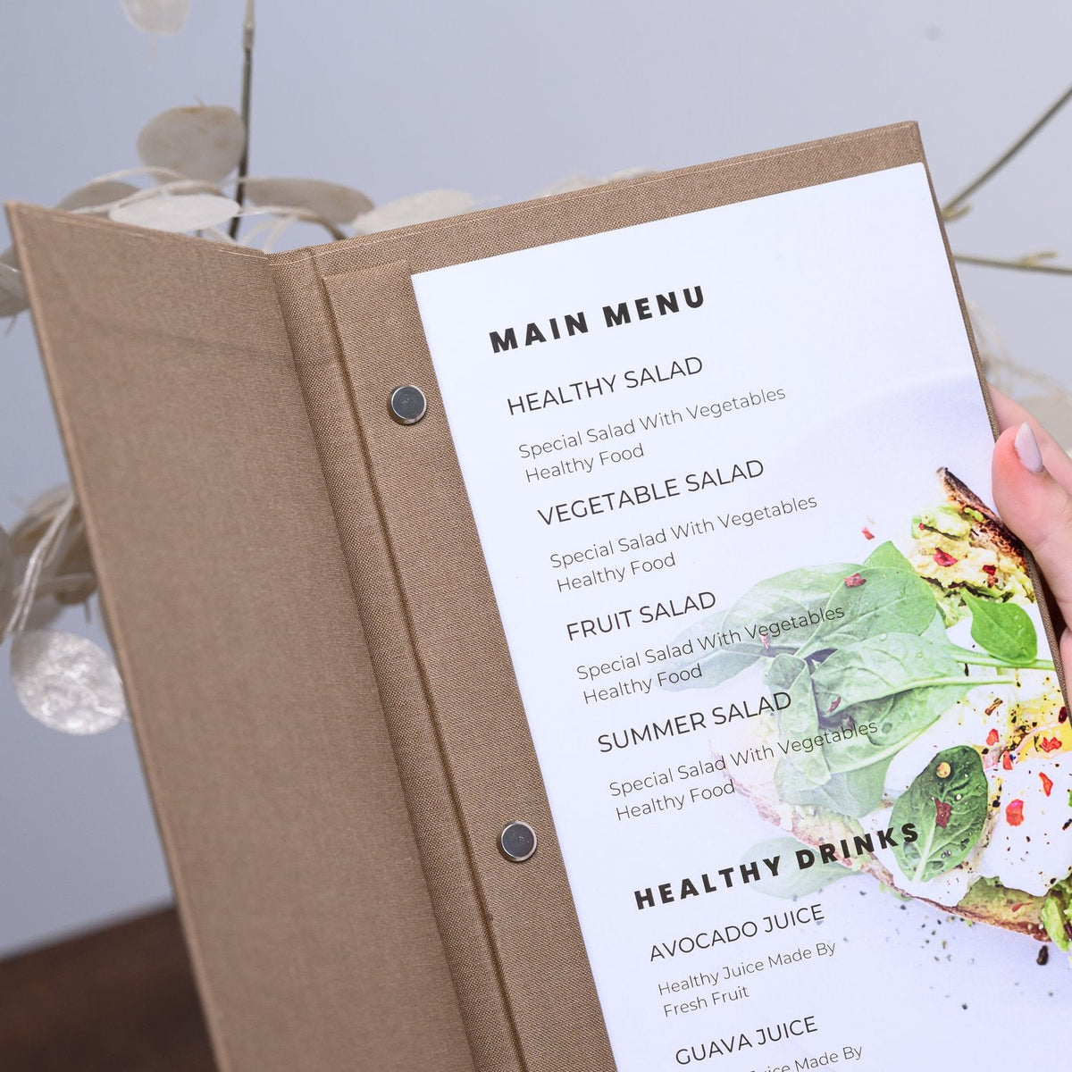 Hand holds an open taupe textile menu folder with a printed menu page and botanical graphic on the table