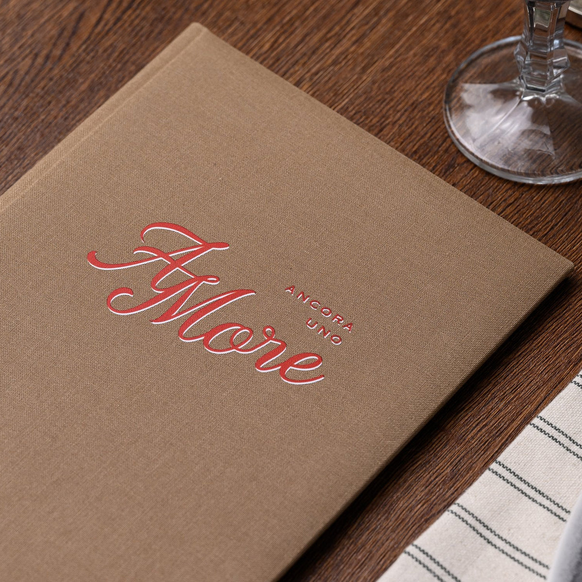 Closed taupe textile menu folder with a red cursive script logo lying on a striped fabric table placemat