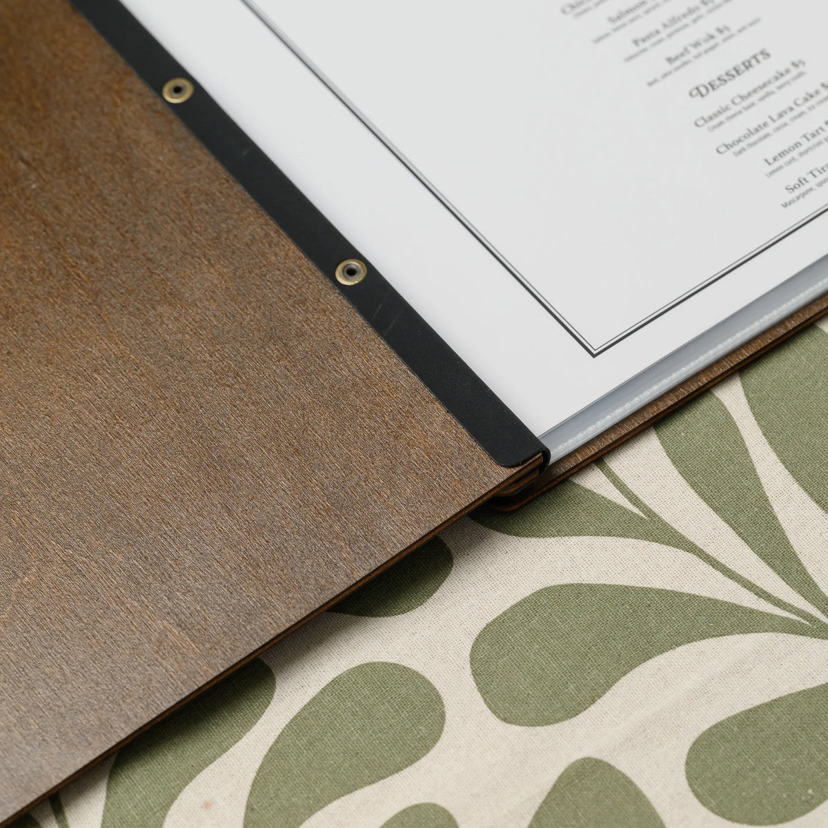 Open walnut plywood menu cover with a printed menu in a clear sleeve held by a black leather strap with eyelets