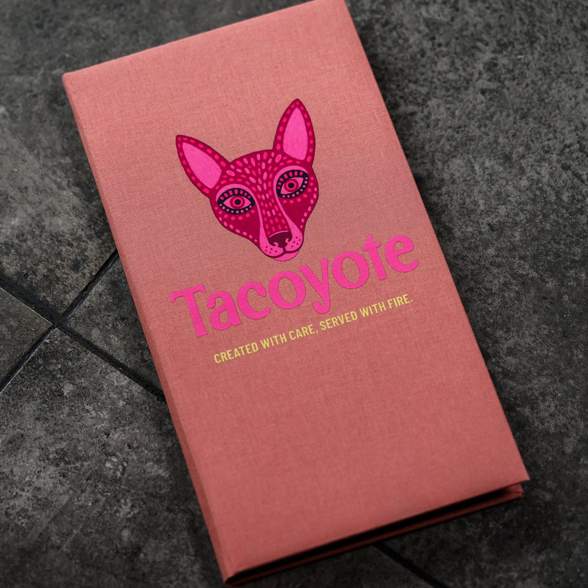 Closed coral-pink textile menu folder standing upright with a fox graphic on a dark grey stone surface