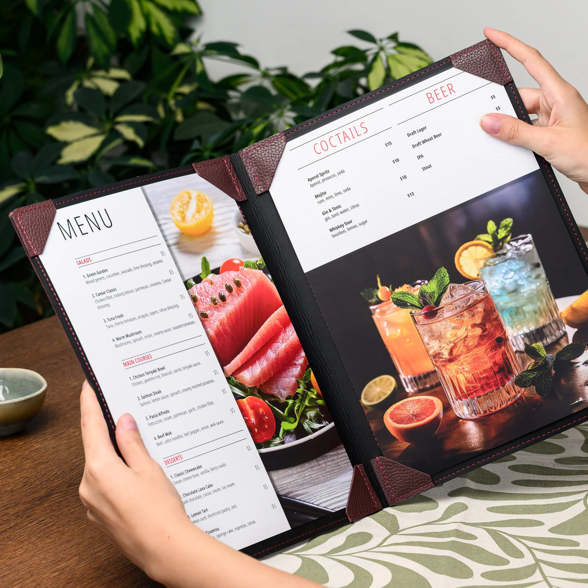 Hands holding an open menu cover with food and cocktail menu sheets secured by bordeaux leather corner tabs
